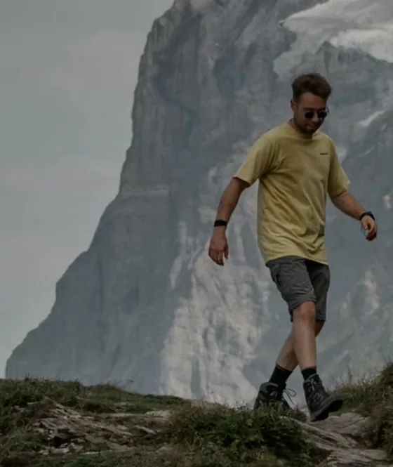 Hiker in a yellow shirt walking down a grassy ridge below a tall rock face